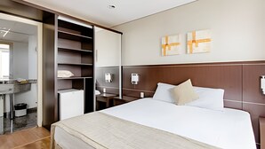 A hotel room with a bed, a desk, a chair, a wardrobe, and a small refrigerator. - Apartment with 1 parking space - UN1209 (Osasco)