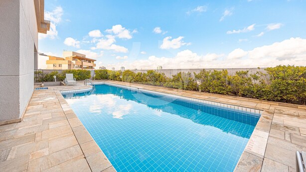 Ein rechteckiger Pool mit Fliesenrand, umgeben von einer Steindeck, mit Blick auf Gebäude im Hintergrund.