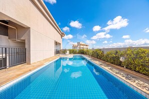 A swimming pool with a tiled edge, a building with a beige facade, and a clear blue sky with clouds. - Apartment with parking space and pool near Alphaville (Osasco)