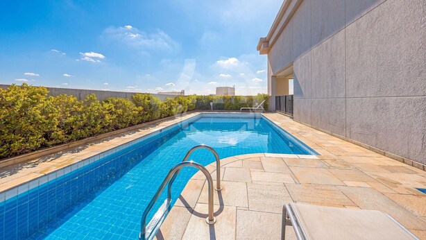 A swimming pool with a metal ladder, surrounded by a stone deck and a low hedge.
