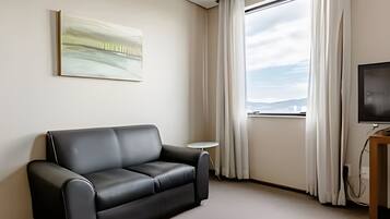 A modern hotel room with a black leather sofa, a small round table, a flat-screen TV, and a large window with curtains.