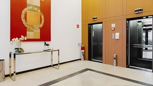 A modern elevator area with a wooden panel wall, a red artwork, and a small table with a vase of flowers. - Apartment with 1 parking space | UN1103 (Osasco)