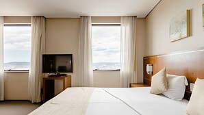A hotel room with a large bed, a TV on a stand, a bedside table, and a view of the city through the windows. - Apartment with 1 parking space | UN1604 (Osasco)