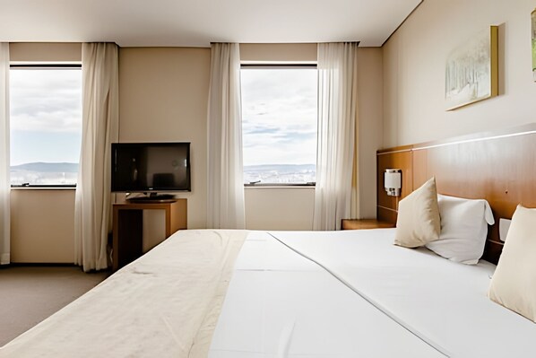 Ein Hotelzimmer mit einem großen Bett, einem Fernseher auf einem Standfuß, einem Nachttisch und Blick auf die Stadt durch die Fenster.