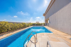 A swimming pool with a metal ladder, surrounded by a stone deck and a low hedge.
