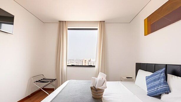 A modern hotel room with a large bed, a nightstand, a window with curtains, and a view of the city. - Apartment with kitchenette - includes 1 parking space - UN1309 (Osasco)