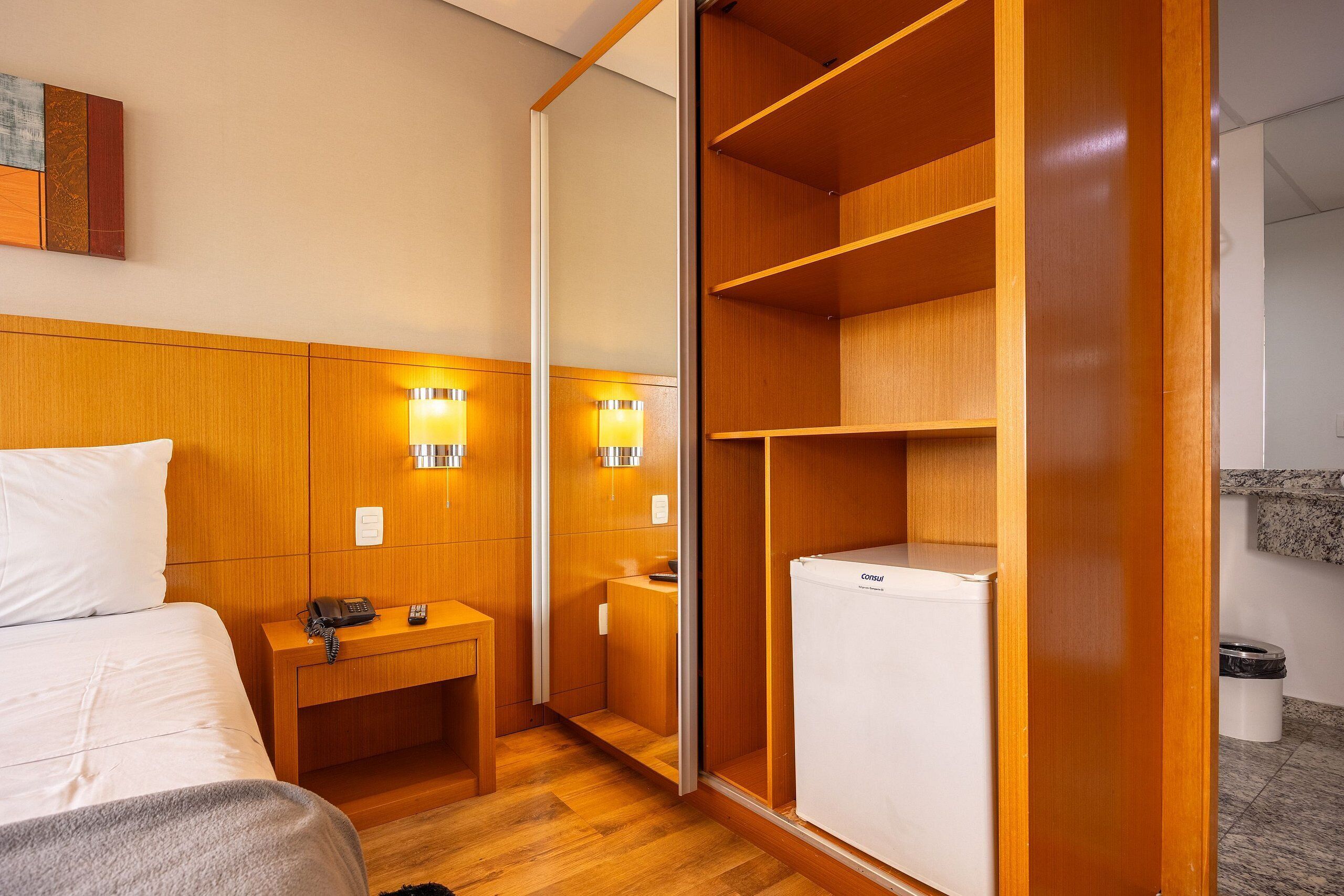 A hotel room with a wooden headboard, bedside table with a phone, a small desk with a lamp, a wardrobe, and a mini-fridge.