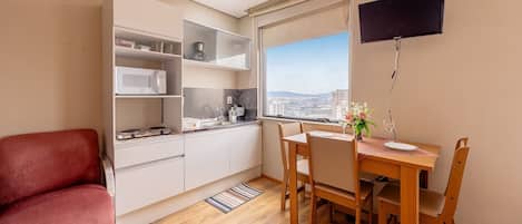 Cocina compacta con armarios blancos, microondas y comedor con mesa y sillas. Se ve una silla roja y un televisor montado en la pared.