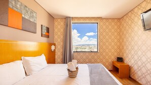 Ein Hotelzimmer mit einem großen Bett, einem hölzernen Kopfteil, einem Fenster mit Blick auf die Stadt, einem kleinen Nachttisch aus Holz und einem an der Wand befestigten Fernseher.