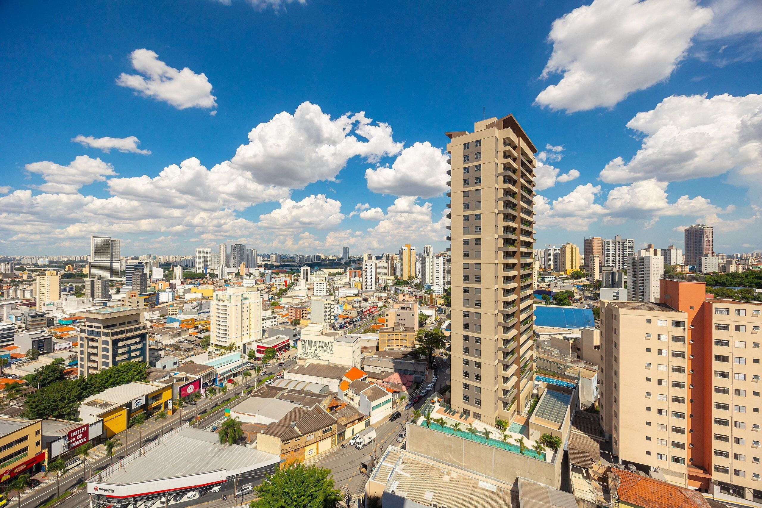 A high-rise building with a modern architectural design, surrounded by a dense urban cityscape with various commercial buildings and a clear blue sky with scattered clouds.