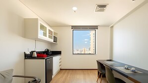 A compact kitchen with a microwave, a coffee maker, and a red container. There is a dining area with a table and chairs, and a window with a city view. - Hospedez - Beautiful apartment near Alphaville ap1505 (Osasco)