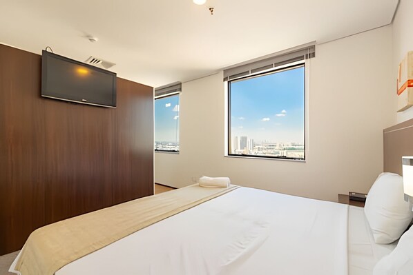 A hotel room with a large bed, a TV mounted on the wall, and a city view through the windows.
