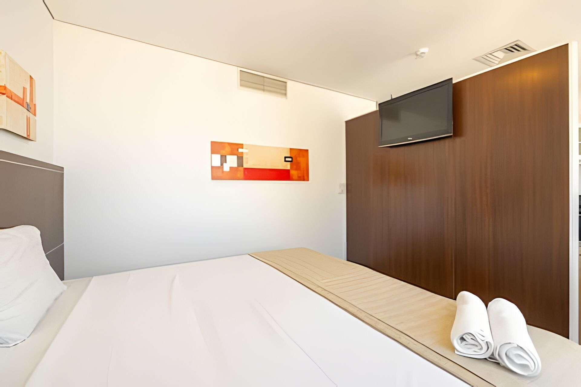A hotel room with a bed, a wooden headboard, a flat-screen TV mounted on the wall, and a small artwork above the bed.
