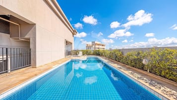 Uma piscina com borda azulejada, um edifício com fachada bege e um céu azul claro com nuvens.