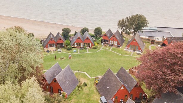 Un hôtel situé sur la côte, avec des petits chalets aux toits rouges disposés en demi-cercle, une pelouse verdoyante et une plage en arrière-plan.