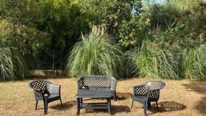 A patio area with a black metal bench and two chairs, surrounded by tall grass and trees.