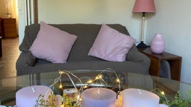 A living room with a glass coffee table, two purple cushions on a grey sofa, a pink lamp, and a painting on the wall.
