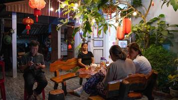 A group of people seated around a wooden table in an indoor setting with hanging plants and lanterns.