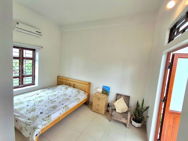 Un dormitorio con una cama individual, una mesita de noche de madera, una silla, una planta en maceta y una ventana con barrotes.