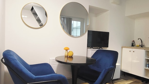Una habitación con dos sillas azules, una mesita redonda con un objeto amarillo, dos espejos en la pared, un televisor y un gabinete blanco con un lavabo.