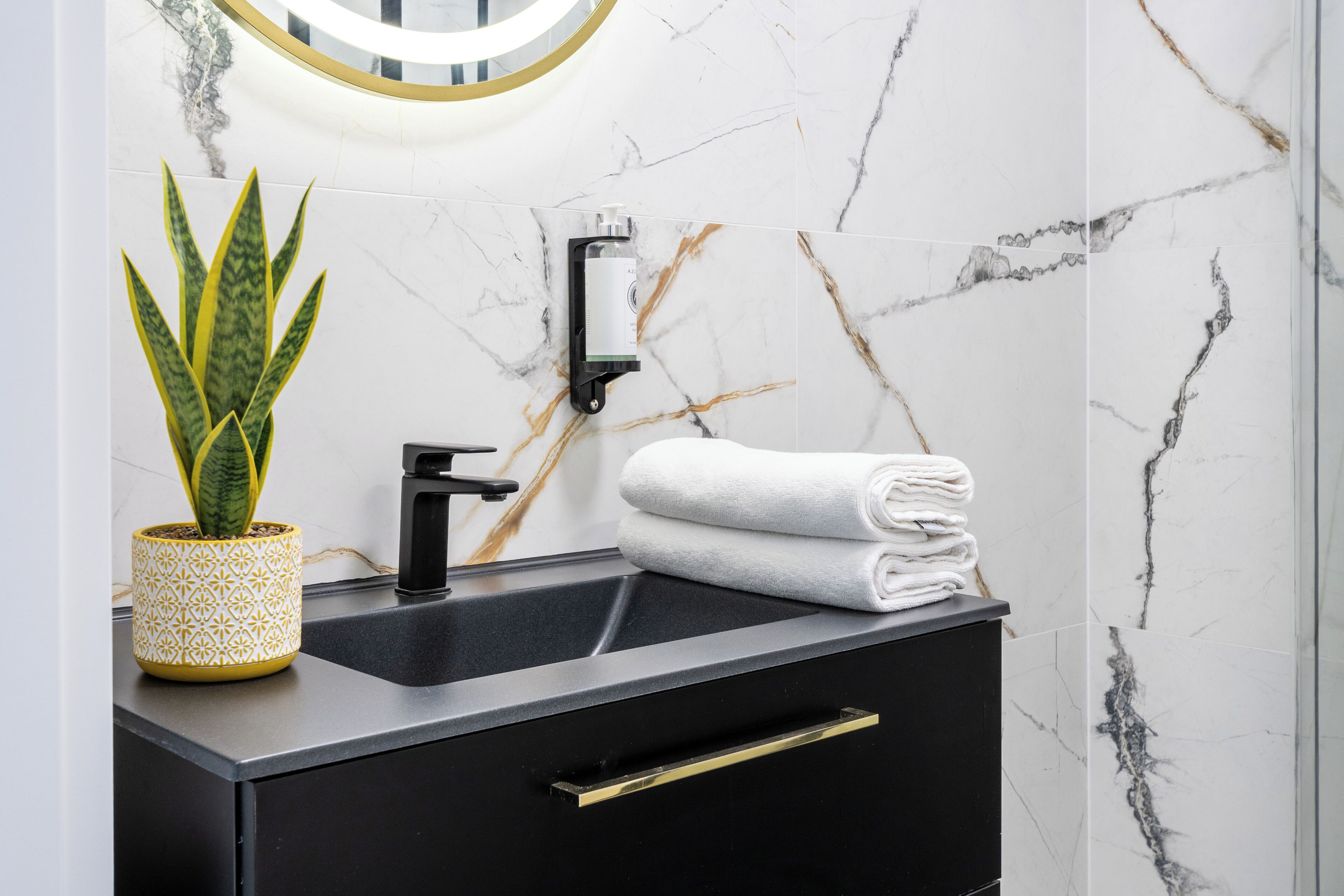 A modern bathroom with a black sink, marble walls, a potted plant, and a towel stack.