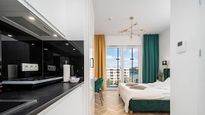 Habitació d'hotel moderna amb zona de cuina, un llit amb capçalera tapissada de verd i vistes a la ciutat a través de grans finestres.