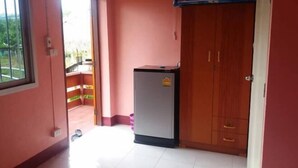 A room with a wooden wardrobe, a small refrigerator, a fan, and a window with a view of greenery. - Khuang Na Farmview (Mae Chaem)