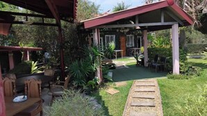 A covered outdoor area with a dining table and chairs, surrounded by lush greenery and a stone pathway. - Khuang Na Farmview (Mae Chaem)