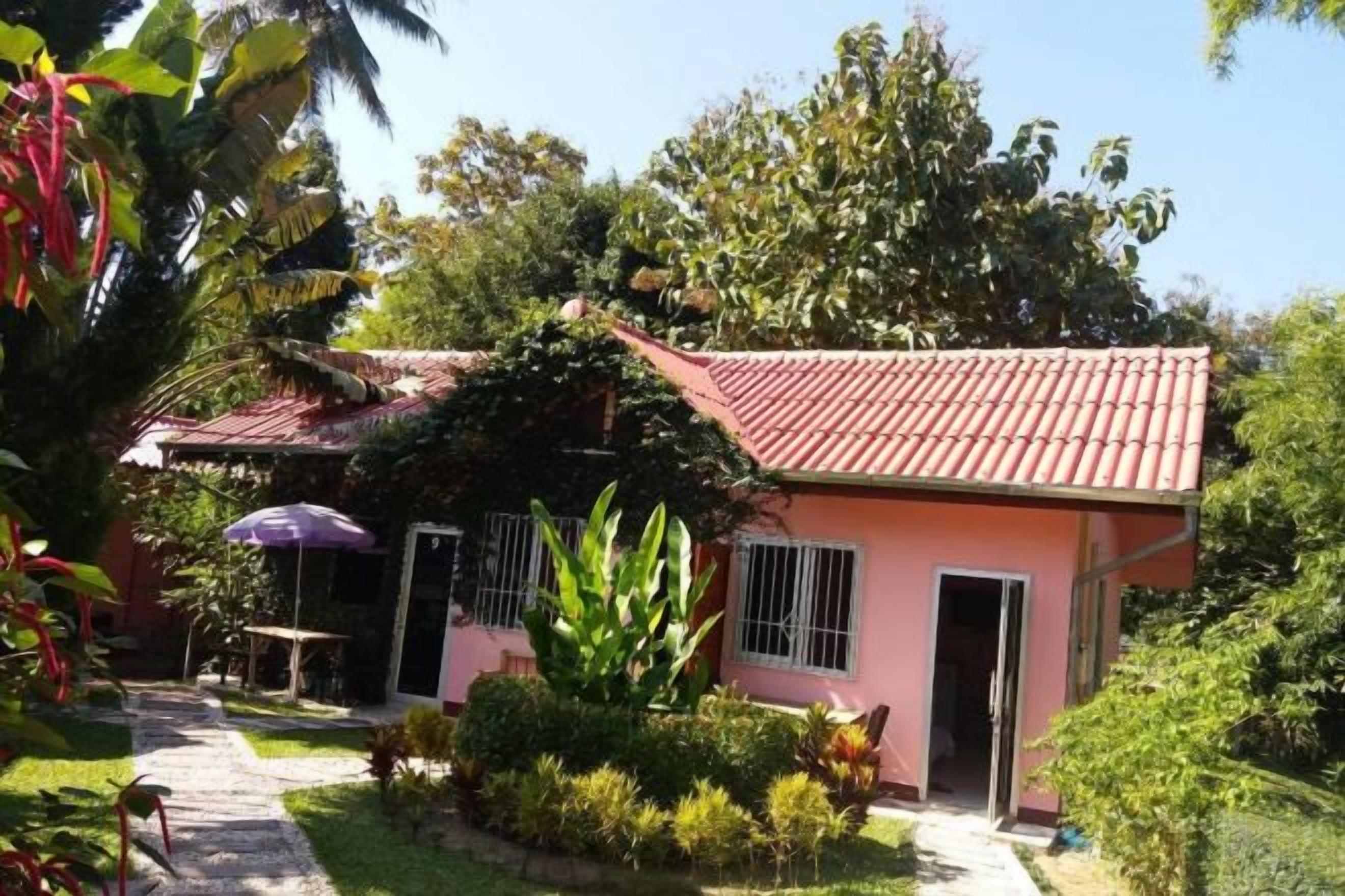 Un edificio rosa con tejado de baldosas, rodeado de follaje y un sendero que lleva a una puerta.