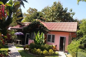 A pink building with a tiled roof, surrounded by greenery and a pathway leading to a door. - Khuang Na Farmview (Mae Chaem)
