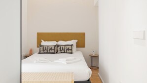 Un lit bien fait, avec une literie blanche, une tête de lit en bois et deux coussins à motifs. On y trouve une petite table de chevet avec une lampe et une télécommande.