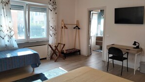 Une chambre à coucher comprenant un lit, un bureau, une chaise, un portant à vêtements et une fenêtre avec des rideaux.