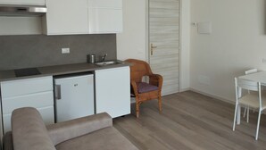 Une petite cuisine équipée avec des meubles blancs, un petit réfrigérateur et un évier. On y trouve une chaise en osier avec un coussin et une table à manger blanche avec une chaise.