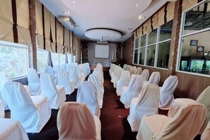 Une salle de conférence avec des rangées de chaises recouvertes d’un tissu blanc et pêche, de grandes fenêtres le long d’un des murs et un écran de projection à l’avant.