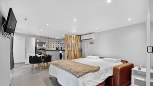 Een moderne hotelkamer met een bed, een bureau, een stoel, een aan de muur bevestigde televisie en een kitchenette op de achtergrond.