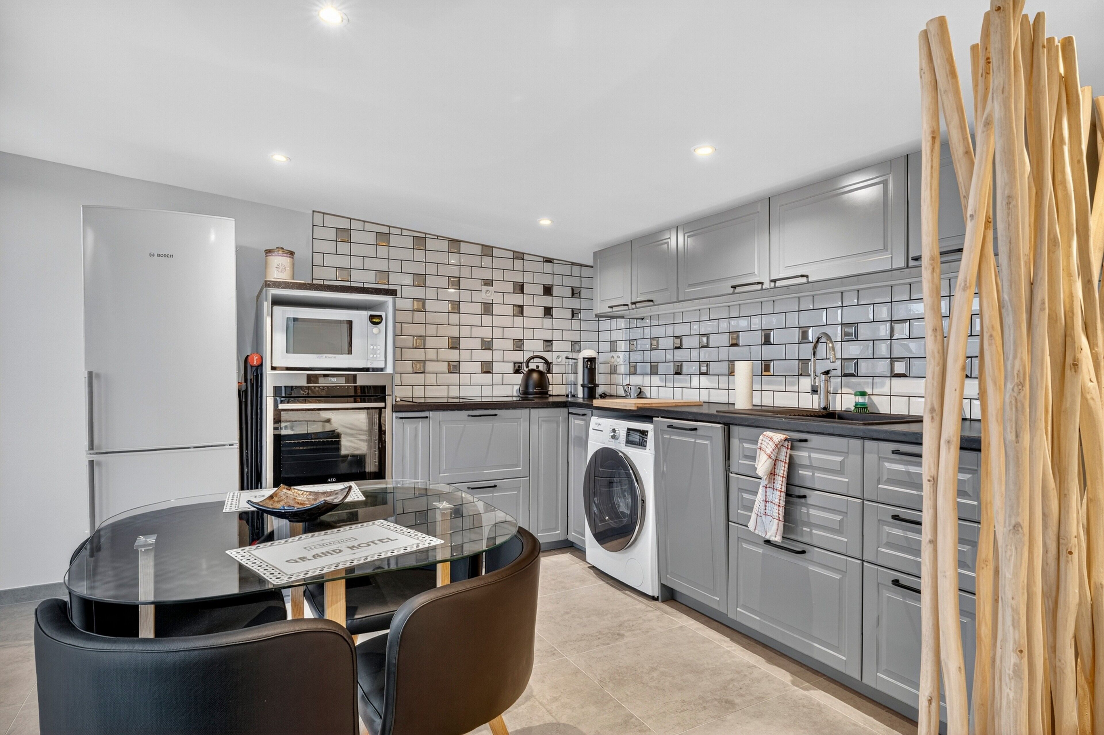 A modern kitchen with grey cabinets, a Bosch refrigerator, a microwave, a washing machine, and a dining area with a glass table and chairs.
