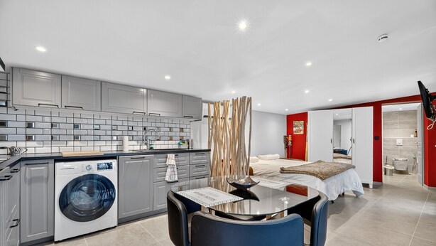 Een moderne keuken met grijze kastjes, een wasmachine en een eethoek met een glazen tafel en blauwe stoelen.