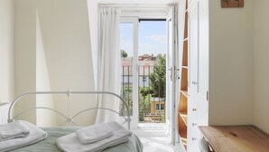 Ein Schlafzimmer mit einem Bett, einem Nachttisch aus Holz, einem Fenster mit Vorhängen und Blick auf Gebäude.