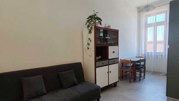 A living room with a dark sofa, a wooden cabinet with glass doors, a dining table with chairs, and a large window with sheer curtains. - LA CASINA Panoramic apartment in the countryside very close to the village (Amelia)
