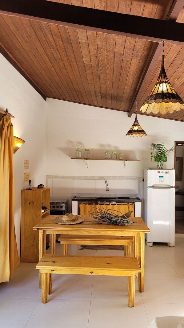 A kitchen with wooden beams, a wooden table with a bench, a stove, a refrigerator, and hanging pendant lights.