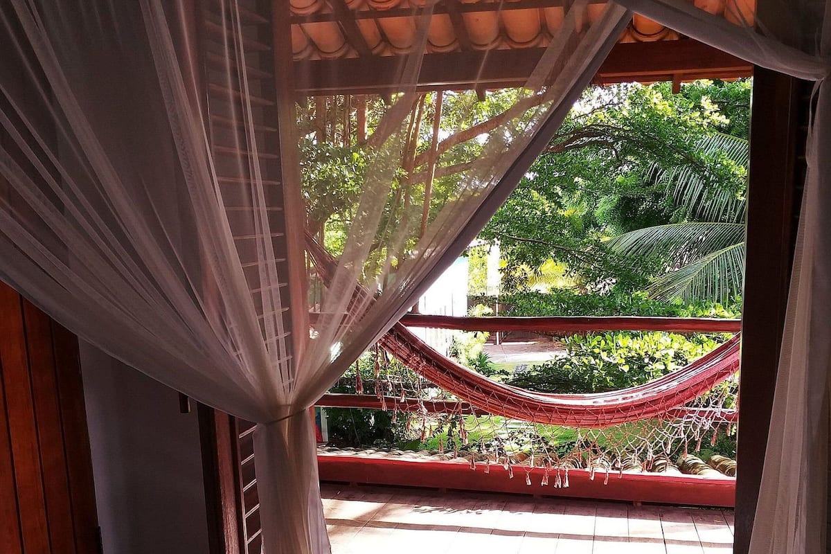 Quarto com cama sob dossel, vista para uma rede e vegetação lá fora.