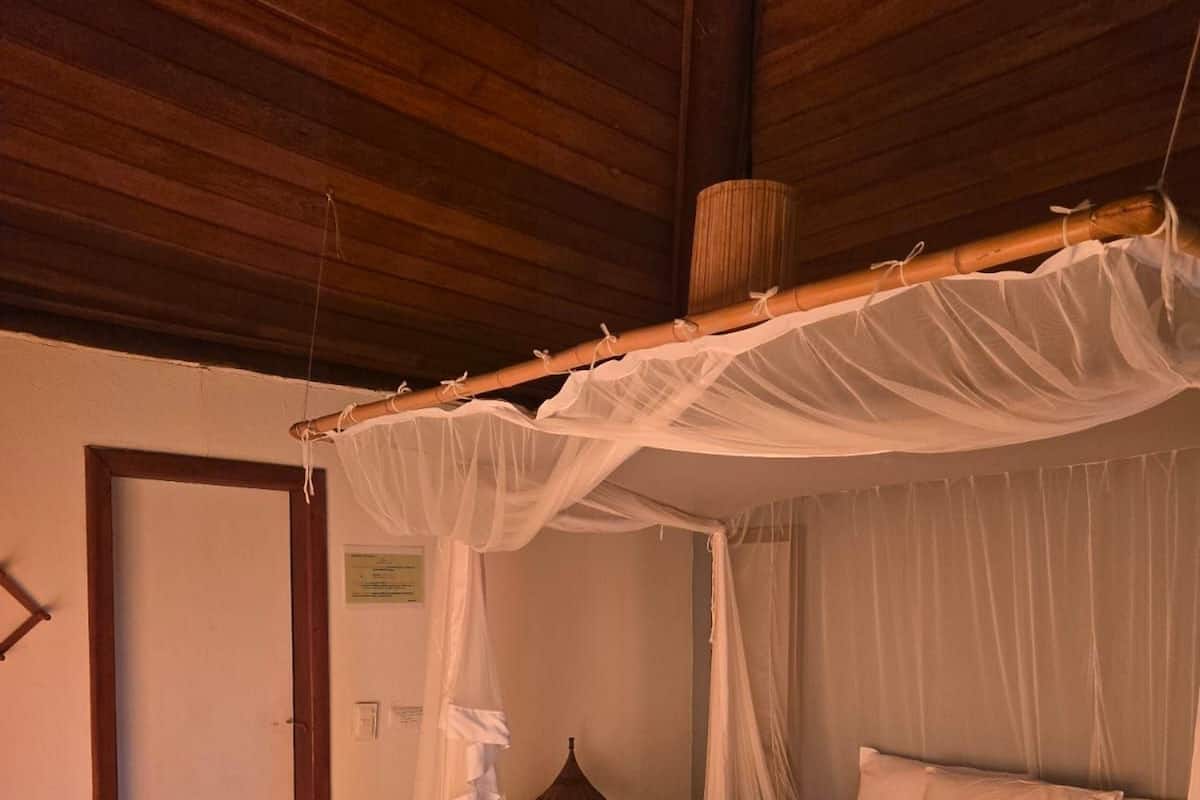 Uma cama de dossel com lençóis brancos, soalho de madeira, uma mesinha e um abajur.