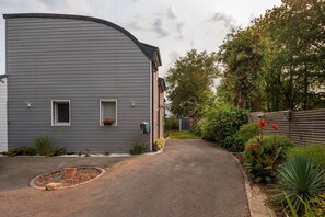 A residential property with a curved grey exterior, a driveway leading to a gated entrance, and a well-maintained garden with various plants and flowers. - Room 'Gwened' with Shared Terrace, Wi-Fi, and Air Conditioning (PLOEREN)