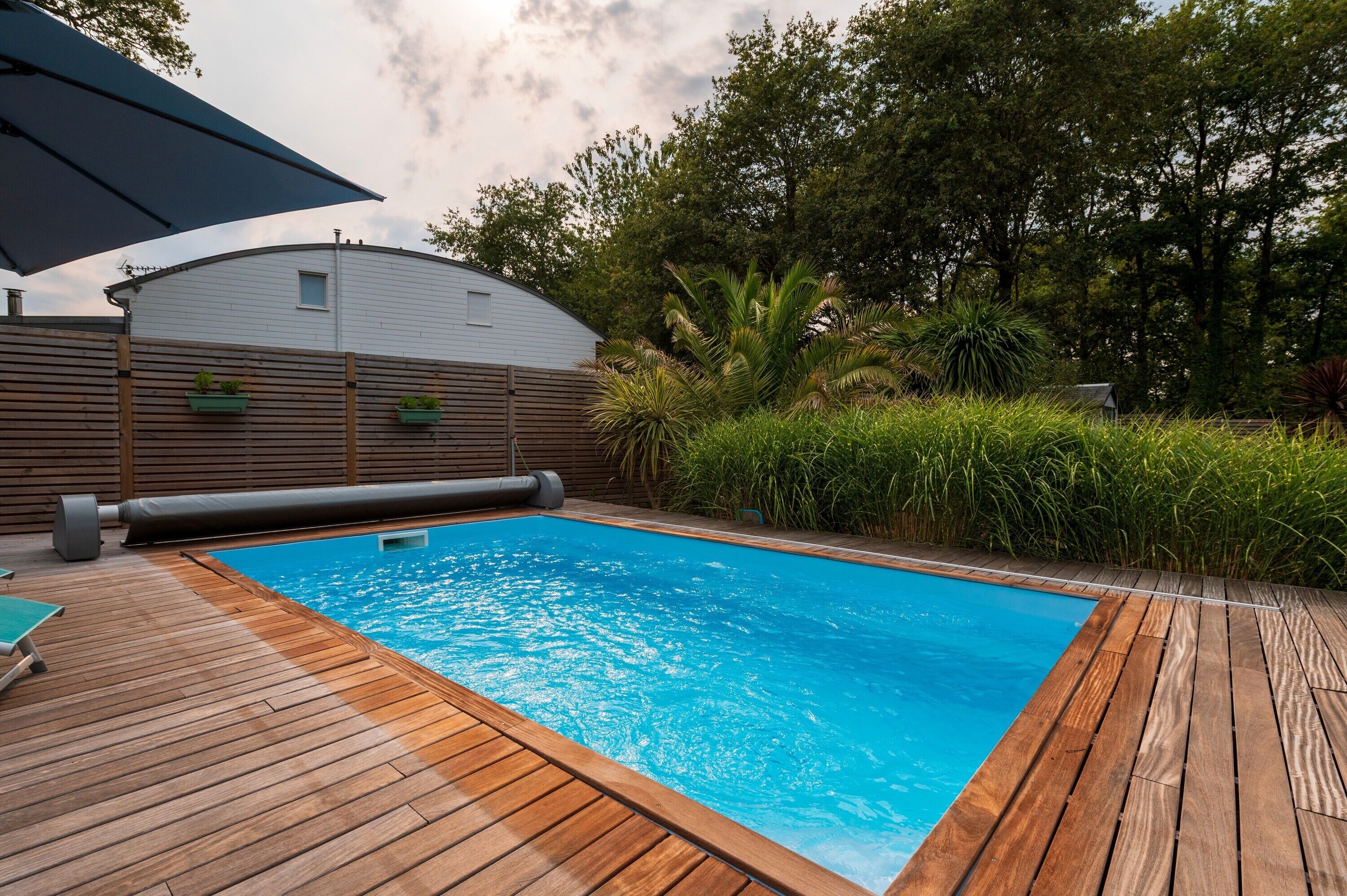 A wooden deck with a swimming pool, surrounded by greenery and a white building.