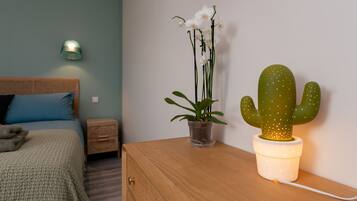A bedroom with a bed, a bedside table with a potted plant, a wall-mounted lamp, and a cactus in a pot.