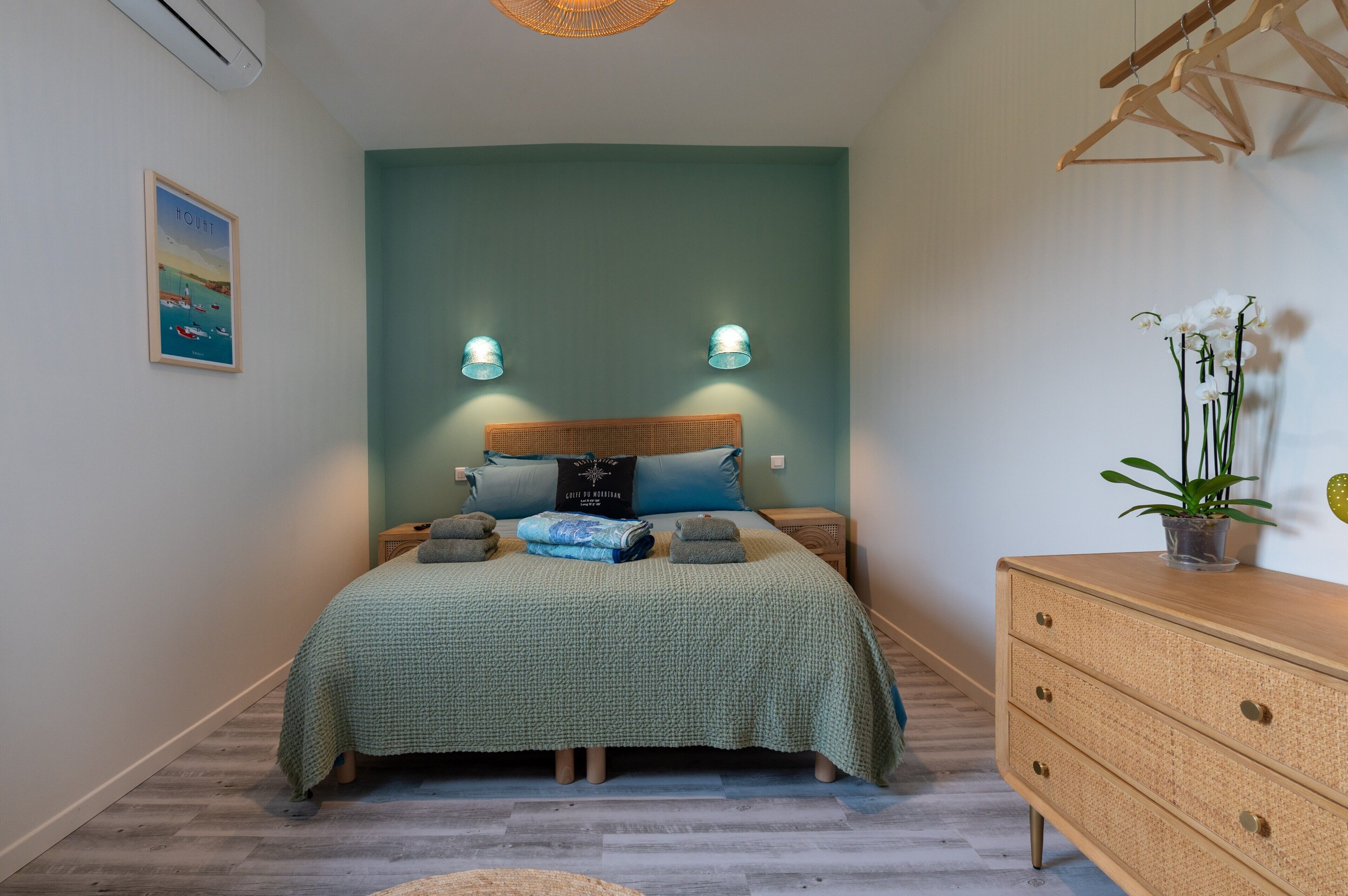 Une chambre à coucher comprenant un lit, des tables de chevet, une commode, un climatiseur fixé au mur, un tableau encadré et un luminaire suspendu.