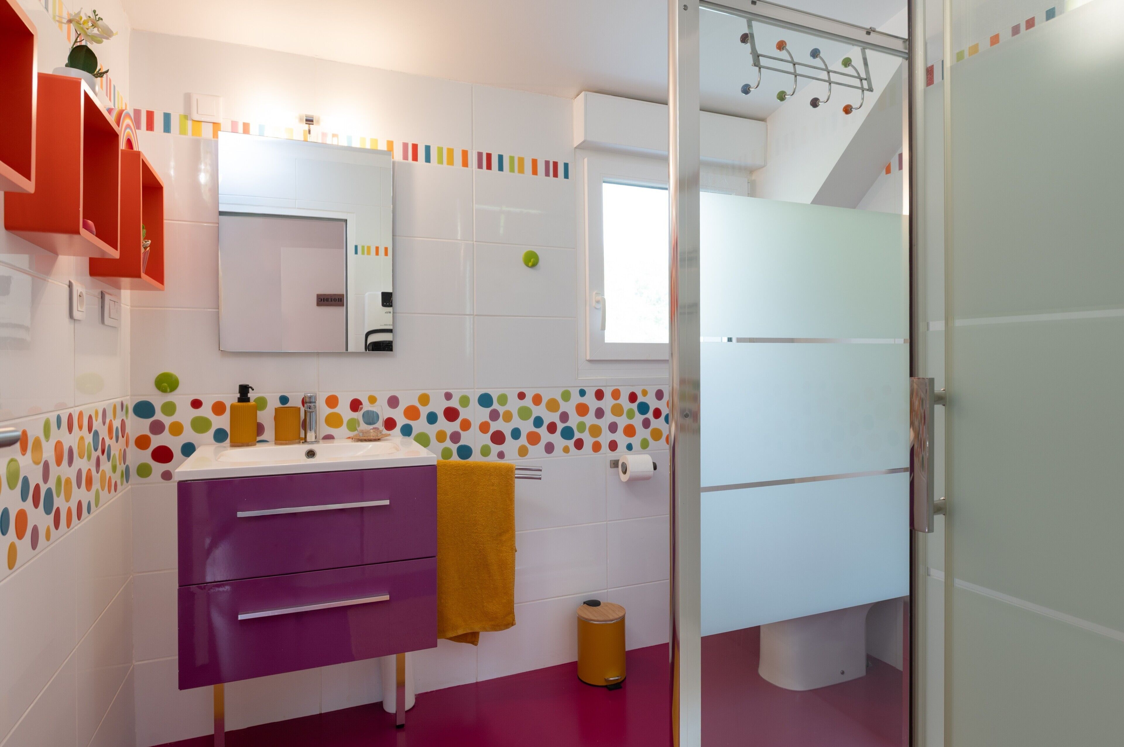 Ein Badezimmer mit einem weißen Waschbecken, einem violetten Schrank und einem Spiegel. Ein Fenster bietet Blick auf eine Treppe.