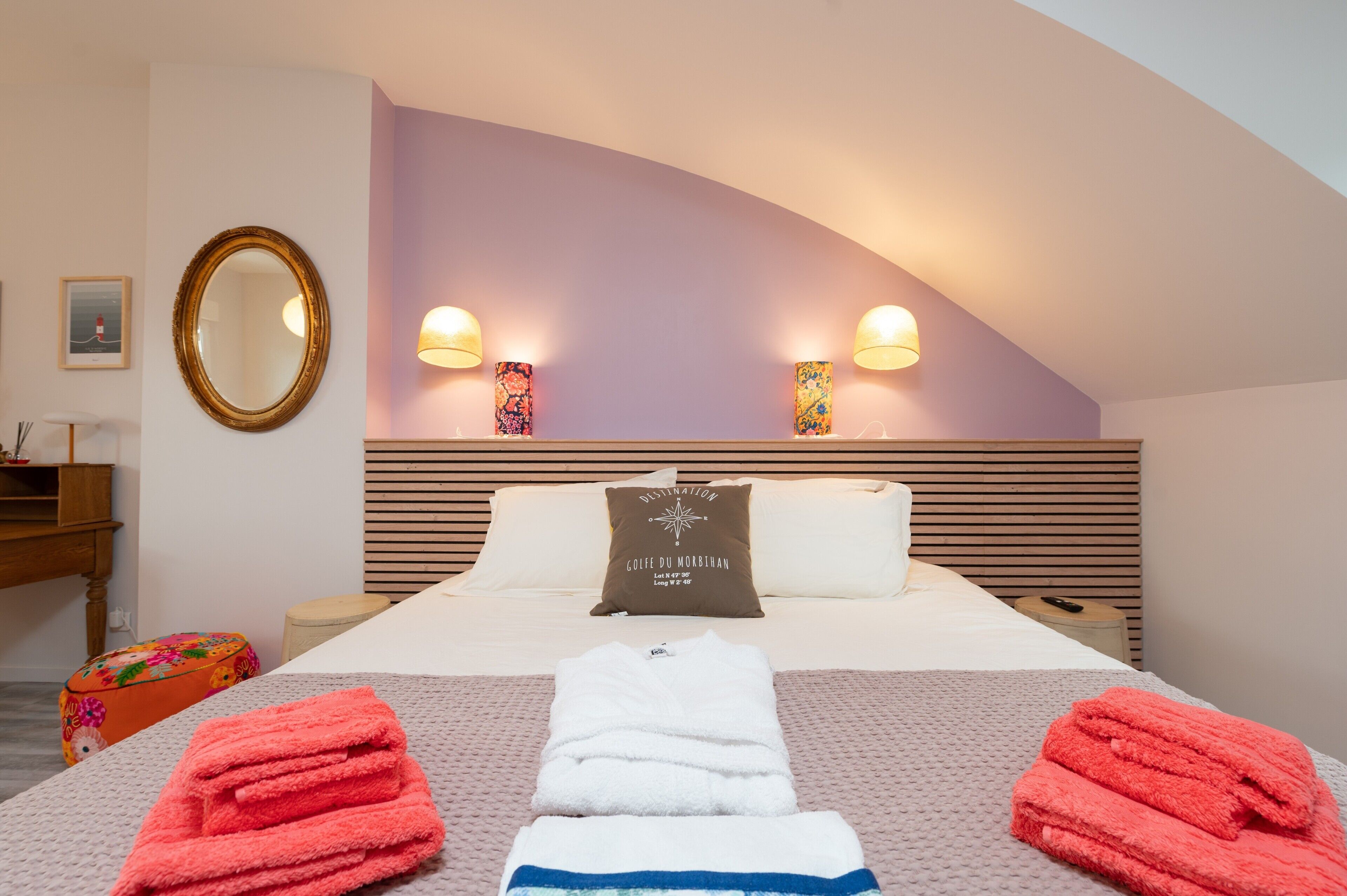 A hotel room with a bed, two wall-mounted lamps, a round mirror, a pillow with a compass design, and a bedside table with a remote control.