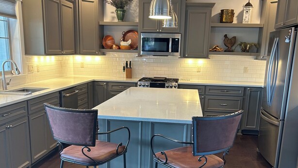 Une cuisine moderne avec des meubles gris, un îlot central doté de tabourets de bar, des appareils en acier inoxydable et un luminaire suspendu.