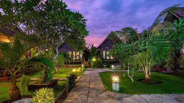 Un sendero conduce a un resort con estructuras de techo de paja, rodeado de exuberante vegetación y céspedes bien cuidados.